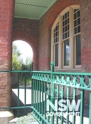 Detail view of the ground-floor residential gate, wrought-iron balustrade and hexagonally-paved porch.  Note the multi-pane arched window to the ground-floor main office, arched brick porch detailing and corrugated iron soffit.