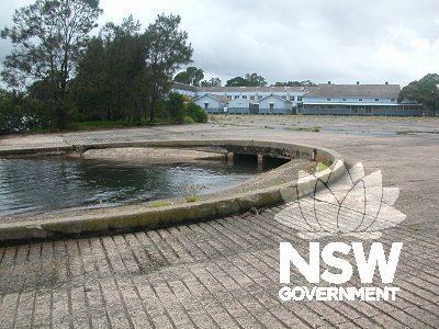 Slipways and stormwater channel in the foreground with part of the original Repair Depot hanger in the background including relocated airmen's sleeping huts, with concrete apron containing remnant aircraft holding rings in the centre.