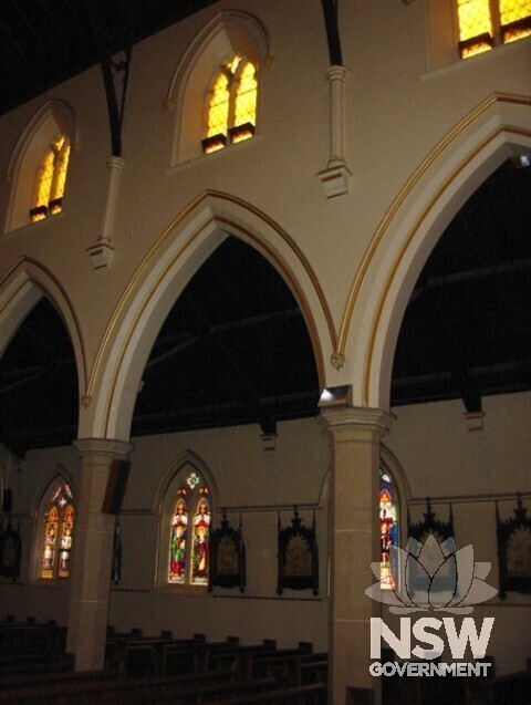 North wall with aisle beyond. Base of columns were once bluestone but are now  ”sanded”.  Stations of the Cross” are visible on the north aisle wall.