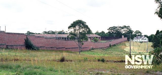 Parts of Ward 21, a disused former maximum security ward, glimpsed across the high north-east wall of the prison, looking west. The photo clearly shows the dip in the ground which allowed the prisoners an outlook to the bush. (Doring Neg.407.25a)